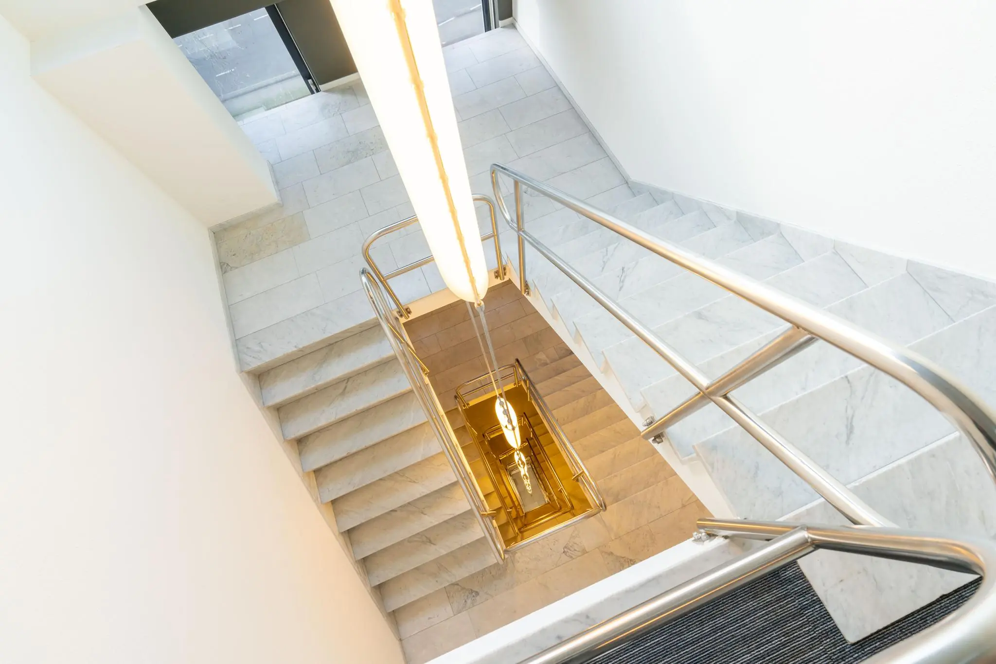 Marmeren trappenhuis met RVS leuningen en modern hanglicht in een gebouw aan de De Bruyn Kopsstraat.